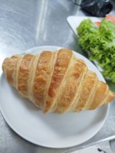 Croissant dorado y crujiente en un plato blanco con ensalada de lechuga fresca al fondo, ideal para desayunos o meriendas.