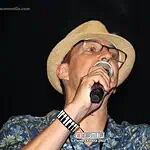 Un hombre cantando en vivo en un concierto, luciendo un sombrero de paja, gafas y camisa de flores, en un escenario oscuro en una fiesta cubana.