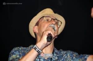 Un hombre cantando en vivo en un concierto, luciendo un sombrero de paja, gafas y camisa de flores, en un escenario oscuro en una fiesta cubana.