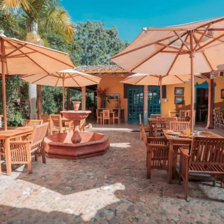 Drinking fountains, a wooden structure, and green plants on the terrace of an open-air restaurant, with umbrellas and wooden furniture in a warm, tropical setting in Colombia.