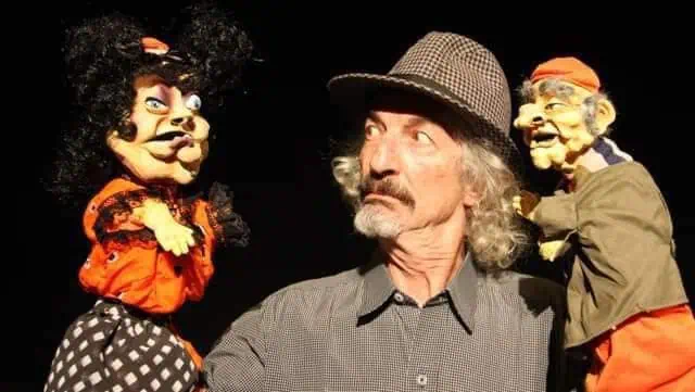 A man with a hat among puppets with funny expressions on a black background, illustrating live events and entertainment for children and adults.