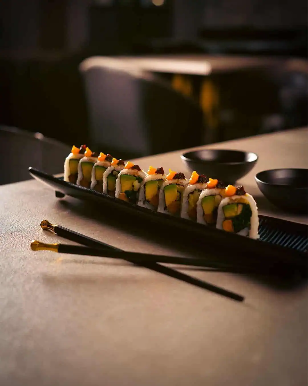 Dünn geschnittenes Sushi auf einem schwarzen Teller mit japanischen Essstäbchen im Vordergrund und schwarzen Schalen im Hintergrund, elegant minimalistischer Stil für Events und Catering in Kolumbien.