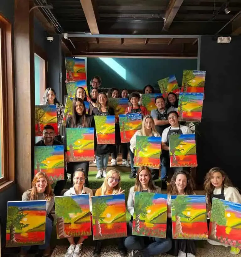 Un groupe coloré de personnes dans un atelier de peinture artistique, exposant leurs œuvres représentant des paysages de coucher de soleil dans l'intérieur chaleureux d'un café ou d'un centre culturel, promouvant la créativité et l'art dans la communauté.
