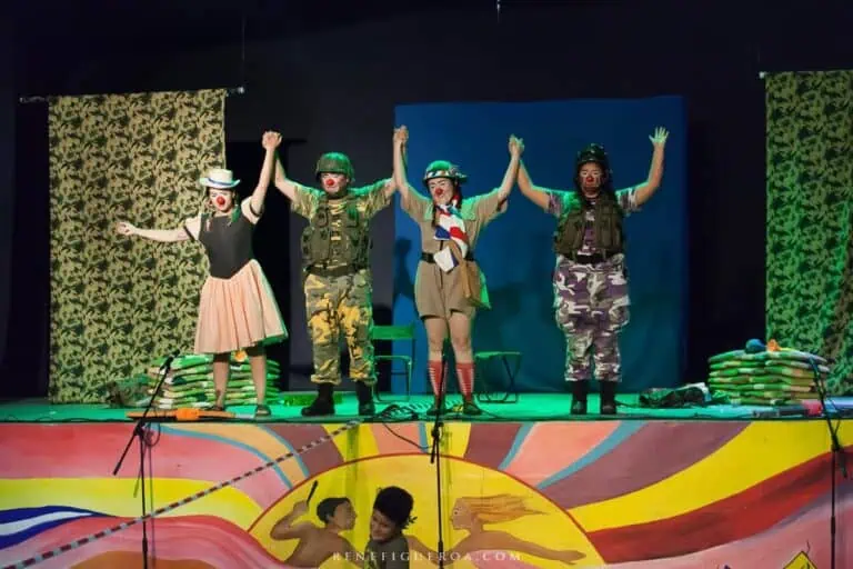 Diversas pessoas vestidas de soldados e palhaços atuam em um palco de teatro, encenando uma peça cômica e teatral com elementos coloridos e acessórios infantis.