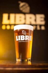 Cerveza artesanal en vaso con logo de LIBRE cervecería, fondo con cartel de LIBRE y cónica de cerveza espumosa.