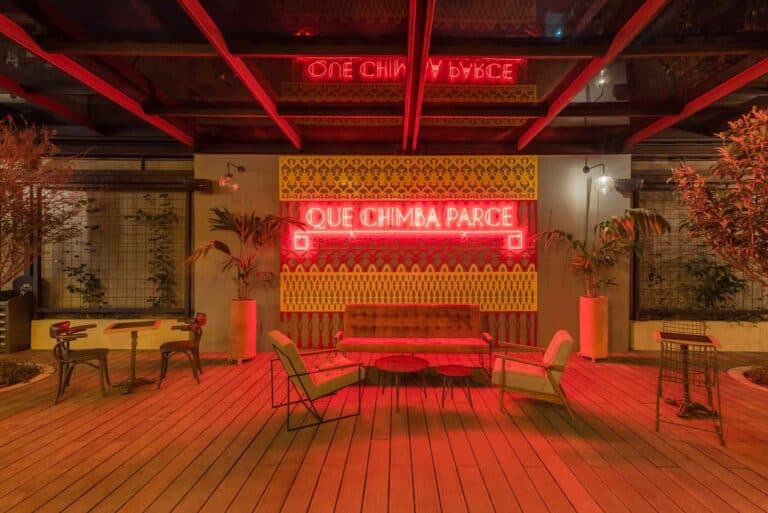 T(er) alt text: Ángulo de la terraza decorada con mobiliario moderno, plantas y luces neón con mensaje en español en fondo de paredes amarillas y borde de madera para eventos y reuniones sociales.