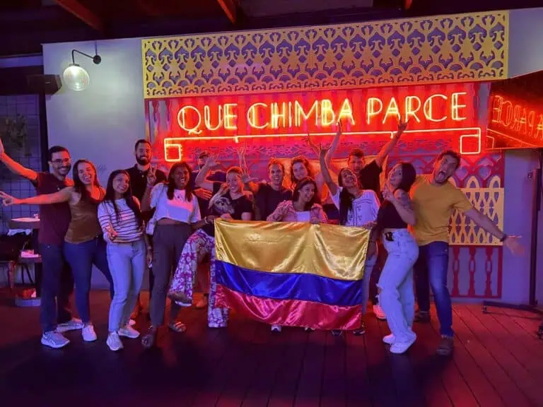 People celebrating with the Colombian flag in front of a neon sign that says "Que Chimba Parce" at a social event.