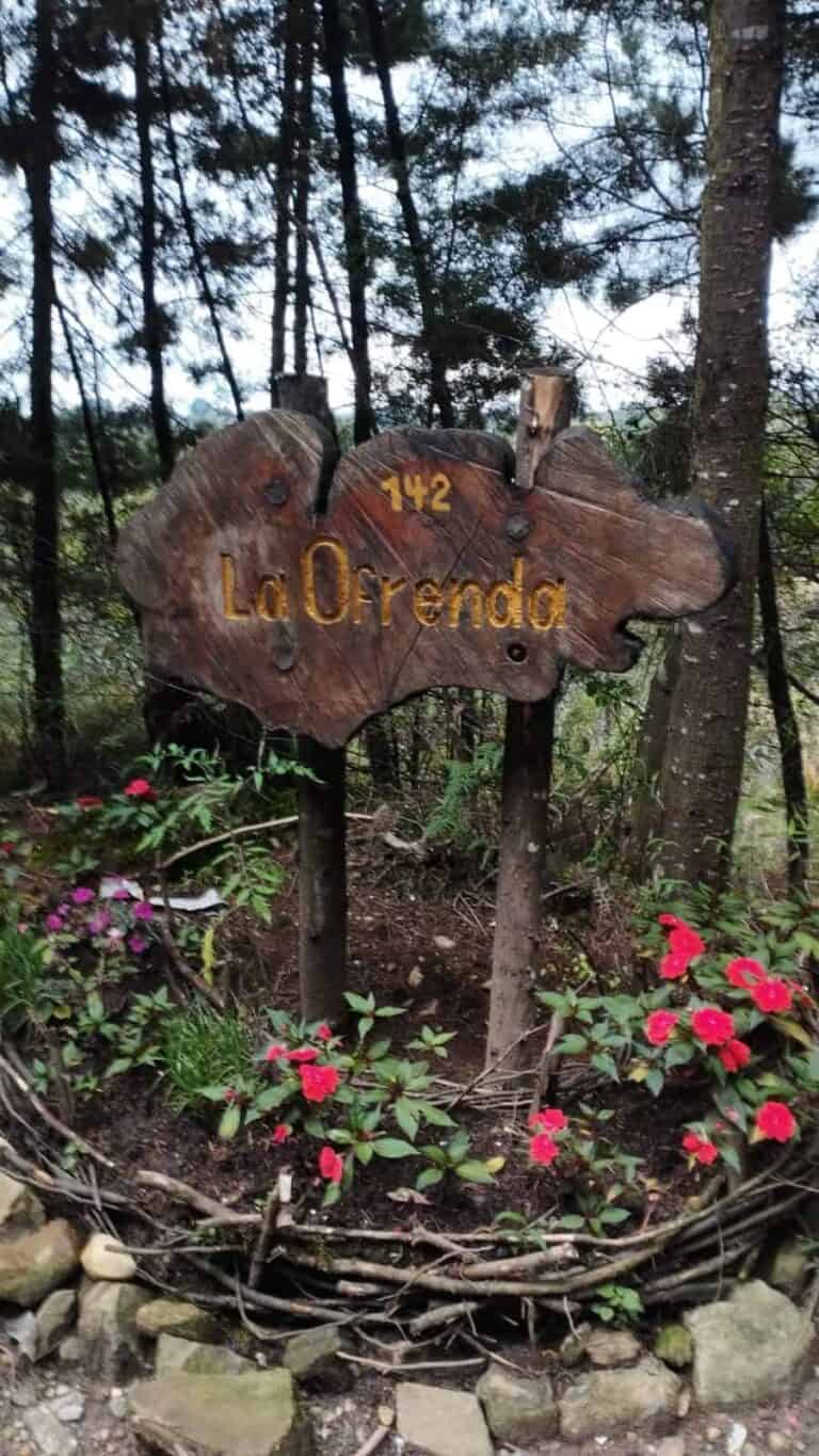 Puerta de madera en un bosque con señal que dice "La Ofrenda" en letras amarillas, rodeada de flores rojas y plantas verdes, indicada para senderismo y turismo ecológico, destino de naturaleza en Colombia.