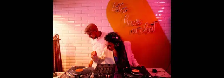 Alt text: Dos DJs en un evento con iluminación rosa y fondo de pared de ladrillos blancos y cartel naranja que dice "Let's have an ad".