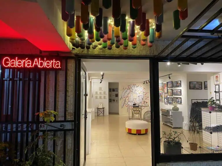 色彩缤纷的艺术装置悬挂在天花板上，艺术画廊入口处有“Galería Abierta”霓虹灯招牌，内部展示着绘画和艺术品，入口附近有植物，现代而充满活力的氛围，哥伦比亚的活动空间。