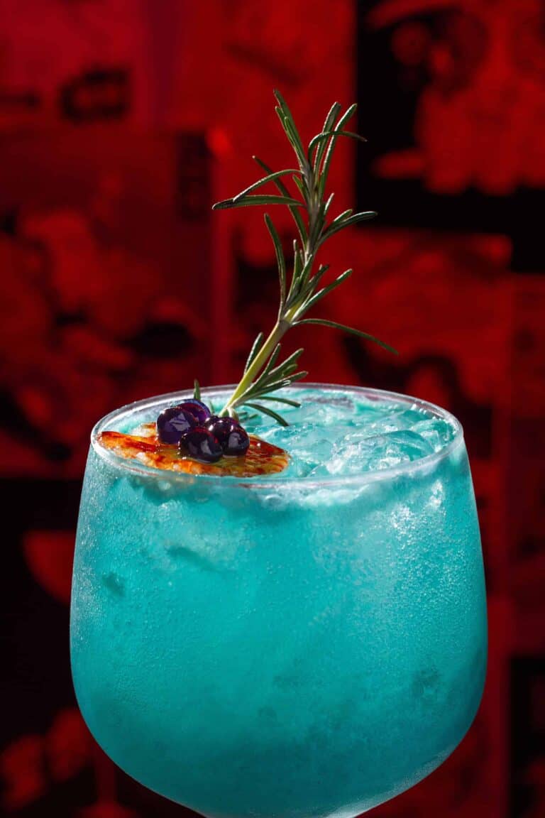 Refresco azul en copa con hielo, decorado con romero, arándanos y una rodaja de fruta tropical, ideal para eventos y celebraciones en Colombia.