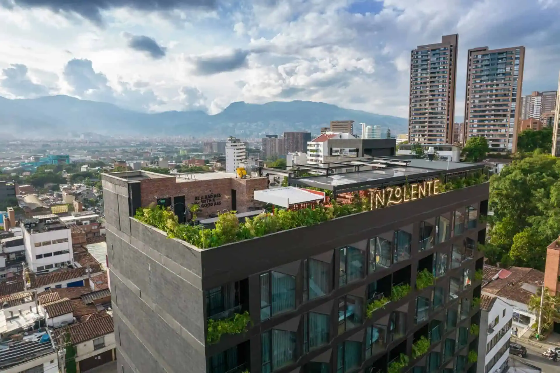 Aerial views of a modern building with a terrace and rooftop garden in an urban setting, with the city skyline and mountains in the background, promoting exclusive events and experiences.