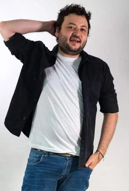 Sonriente hombre joven con barba, vestido informal, posando con actitud relajada en estudio para imagen de evento digital en Colombia.