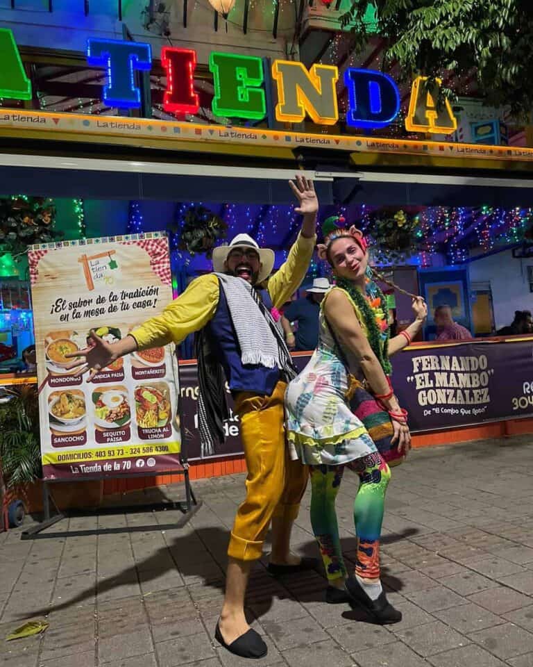 Colorido cartel y personas disfrazadas celebrando en una feria o evento cultural en exteriores, con luces y decoración navideña, promoviendo la gastronomía y tradiciones colombianas en un ambiente festivo y alegre.
