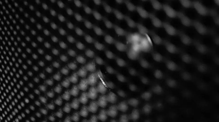 Nahaufnahme eines Metallgewebes, die auf einen bestimmten Bereich fokussiert ist, um Textur- und Musterdetails sichtbar zu machen. Das monochrome Bild hebt die komplexe Struktur des Materials hervor.