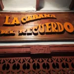 Letrero de madera con texto en relieve que dice "LA CABAÑA DEL RECUERDO" en color naranja.