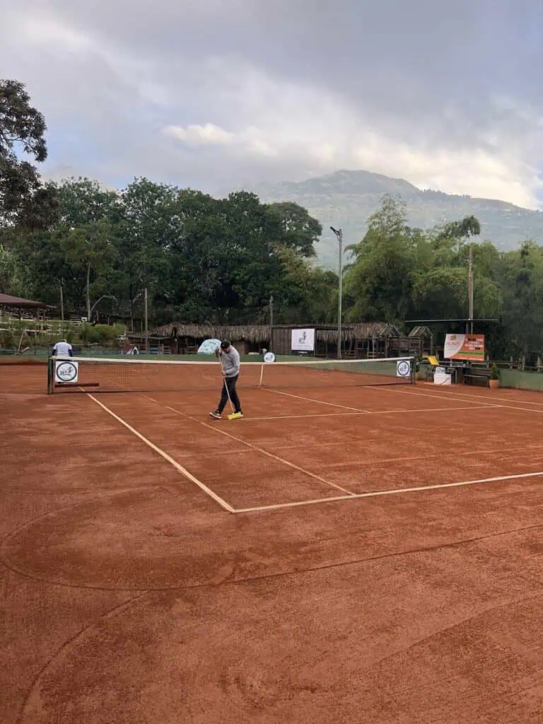 Clay tennis court in a natural setting with mountains and trees in the background, where the field is being maintained by a worker with a sweeper.