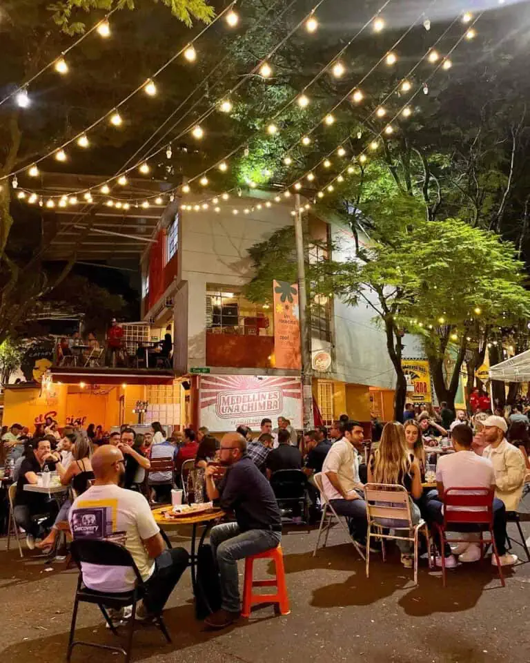 A sky with decorative lights at an outdoor event in Medellín. A festive and social atmosphere with many people enjoying food and music in an urban space with trees and light decorations.