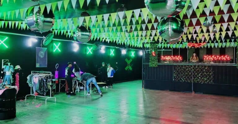 Decoración para evento en interior con banderines verdes, espejos disco y señalización de neon en color rojo, creando ambiente festivo y moderno.