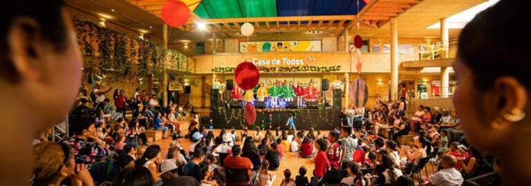 Celebración familiar en espacio cerrado con escenario, niños y adultos disfrutando de entretenimiento, decoración colorida y ambiente festivo en Casa de Todos.