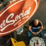 The sign for El Social Tienda Mixta, a traditional establishment since 1969, in an urban setting, features a seated man using his mobile phone, reflecting the local culture and community at social and commercial events.