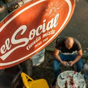 The sign for El Social Tienda Mixta, a traditional establishment since 1969, in an urban setting, features a seated man using his mobile phone, reflecting the local culture and community at social and commercial events.