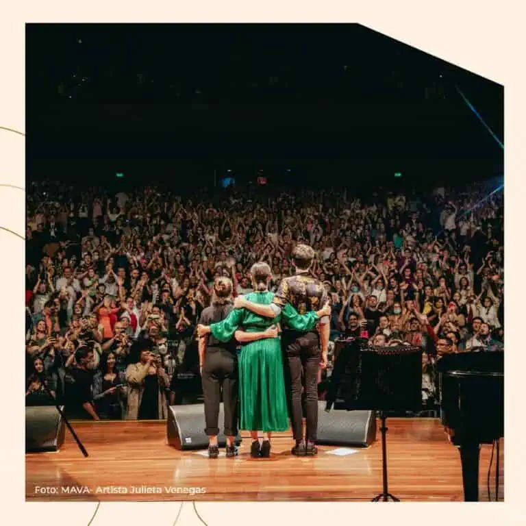 Un groupe d'artistes sur scène disant au revoir au public lors d'un événement musical, avec un public nombreux et passionné, lors d'un concert dans un auditorium.