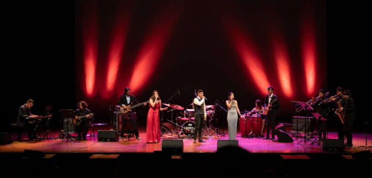 Conjunto de músicos tocando en un escenario iluminado con luz roja, presentación en vivo de bandas y artistas, evento musical, entretenimiento y cultura en un teatro o auditorio.