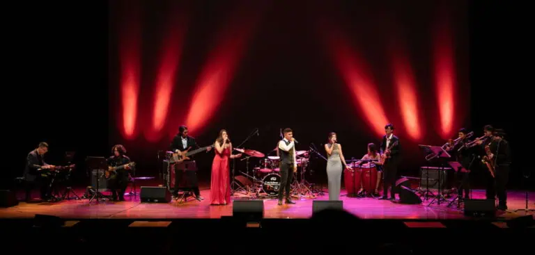 A group of musicians playing on a red-lit stage; a live performance by bands and artists; a musical event, entertainment, and cultural event in a theater or auditorium.
