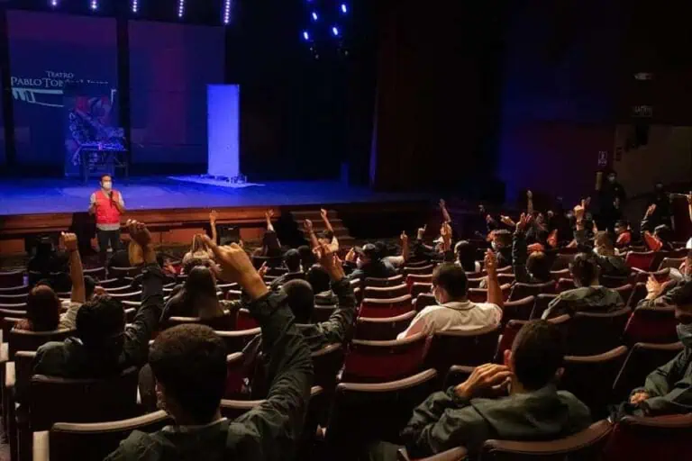 在一个拥有现代灯光和舞台的剧院里，一群年轻的观众在会议或活动中举手发言。