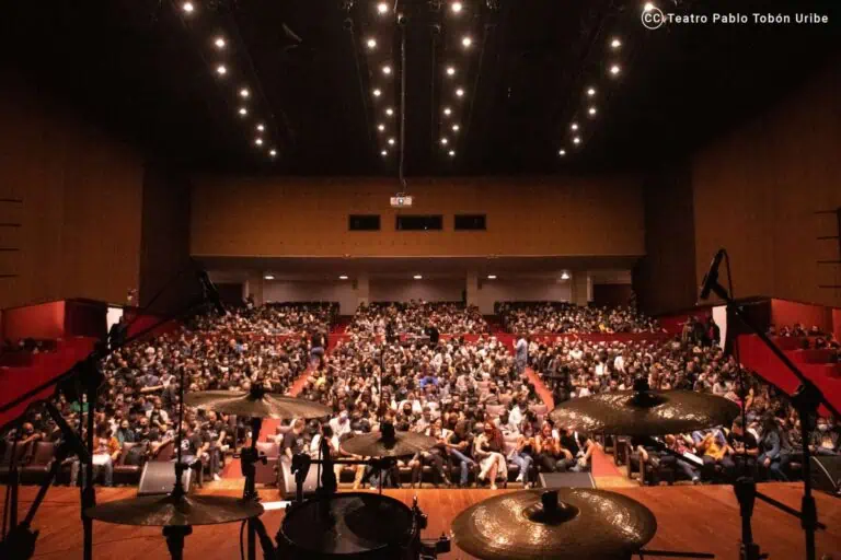 Un auditorium bondé lors d'un événement au Théâtre Pablo Tobón Uribe, préparé pour un concert ou une performance en direct, avec une scène entièrement équipée et un bureau pour les artistes.