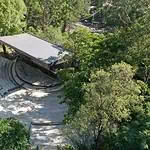 A spacious open-air amphitheater surrounded by nature, ideal for events and concerts in natural settings, with a modern structure and access through lush green areas.