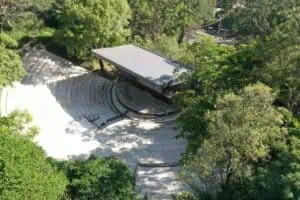 A spacious open-air amphitheater surrounded by nature, ideal for events and concerts in natural settings, with a modern structure and access through lush green areas.