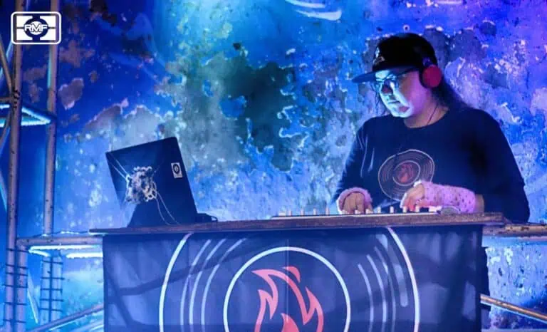 Michael, wearing a black suit with the event logo, blends into the rock wall at an electronic music event, operating his DJ console with modern, portable equipment, in an environment lit with blue lights and vibrant ambiance.