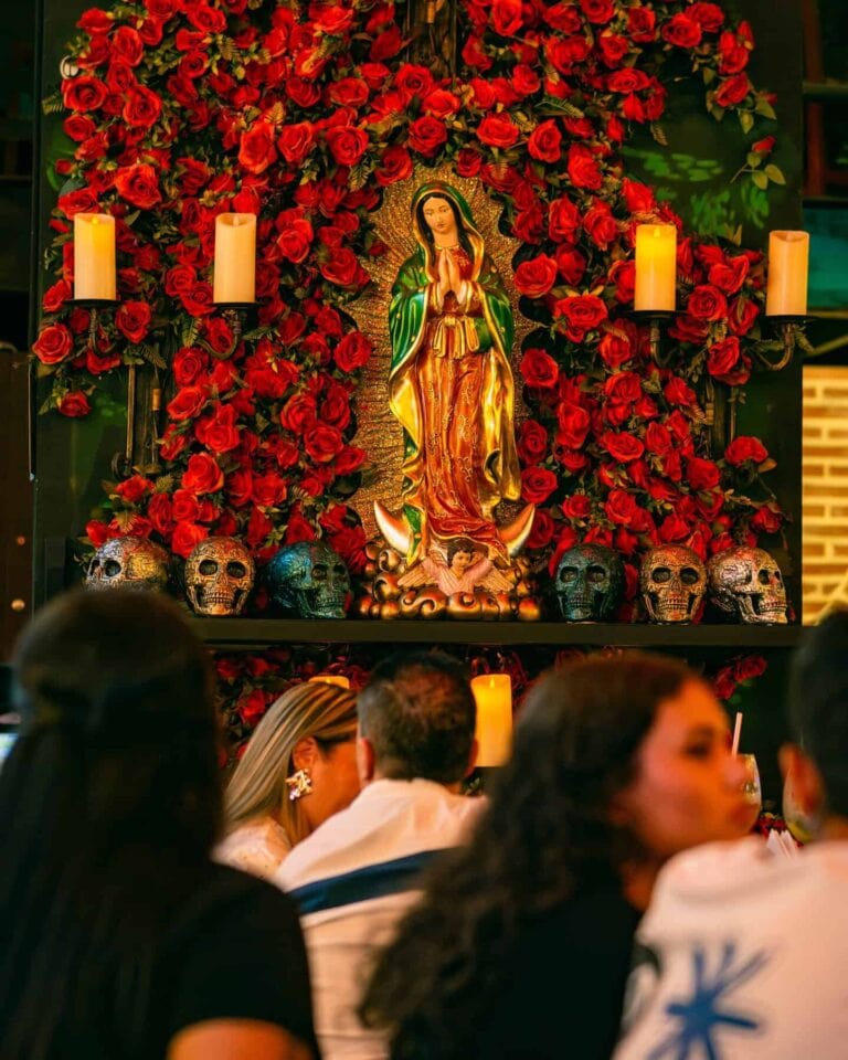 Altar con imagen de la Virgen María rodeada de rosas rojas, velas y calaveras decorativas en un ambiente religioso y cultural.