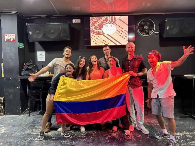 Celebración de grupo con bandera de Colombia en ambiente festivo, mostrando alegría y diversidad en evento social en bar o sala de conciertos. Participantes felices disfrutando la ocasión.