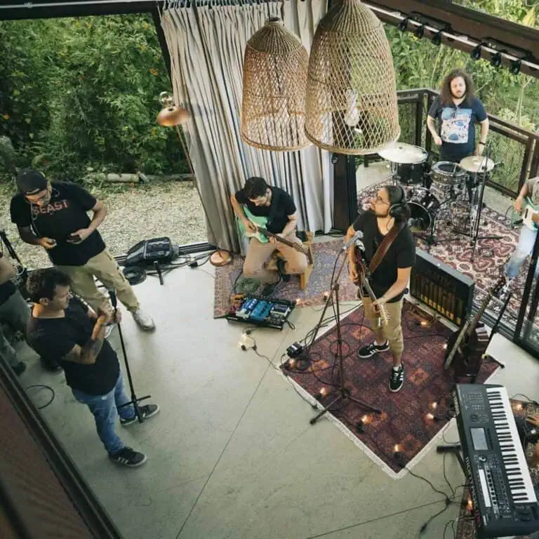 Un groupe musical jouant lors d'un événement en plein air avec des instruments tels que guitare, batterie, clavier et microphones, dans un cadre moderne et accueillant avec un décor bohème et beaucoup de végétation naturelle.