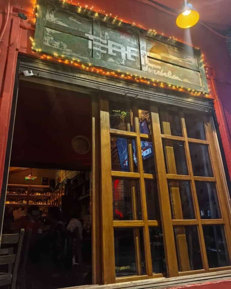 Puerta de madera con estructura de ventanas y cartel de madera con luces decorativas en la fachada de un bar o restaurante, ambientada para eventos, con interior visible y ambiente acogedor.