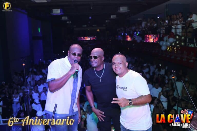 Panel de tres hombres en concierto en un escenario con público, celebración del 6º aniversario La Clave Salsa Bar Medellín en un ambiente festivo y vibrante, con miembros del público disfrutando de la música en vivo.