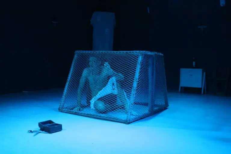 剧院舞台上，一个被关在铁丝笼里的奇怪身影，伴随着蓝色的灯光、昏暗的装饰和背景物件，营造出一种戏剧性和艺术性的氛围，适合文化活动和演出。