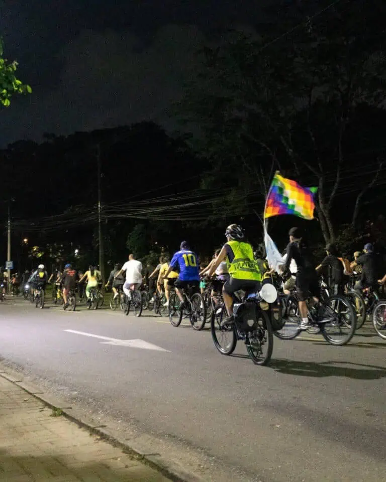 Ciclistas participan en una marcha nocturna por los derechos LGBTQ+ en la ciudad, con banderas de arcoíris, voluntarios y apoyo comunitario en un evento inclusivo y festivo.