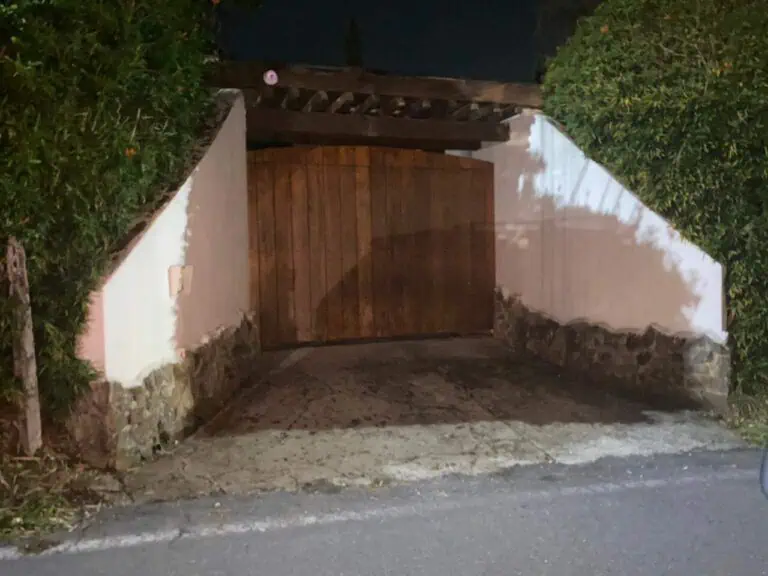 Portail d'entrée en bois de style rustique entouré de murs et de végétation, avec un éclairage nocturne qui met en valeur son design dans un cadre extérieur.
