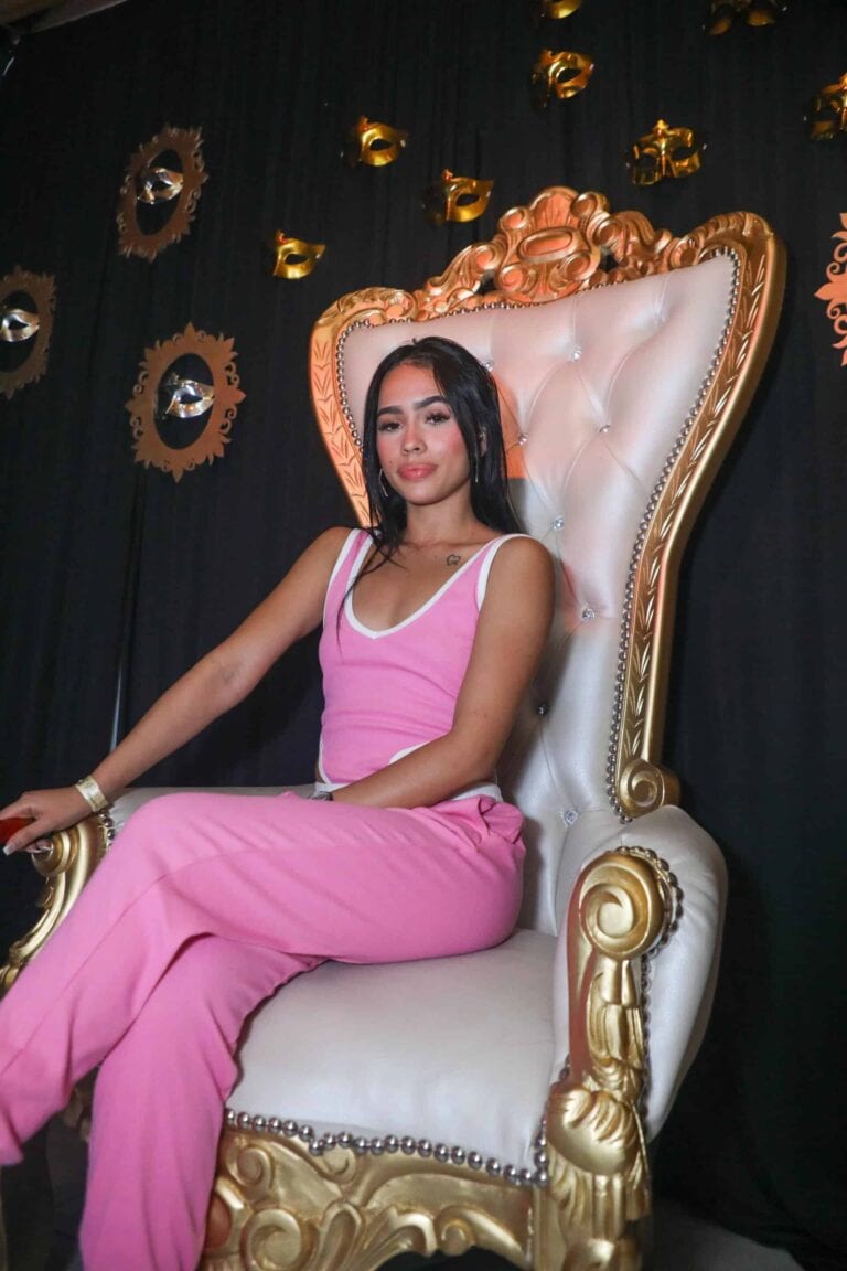 Reina de evento en trono dorado con fondo negro y decoraciones de máscaras doradas, luciendo un conjunto rosa y accesorios elegantes, ideal para celebración y eventos especiales en Colombia.