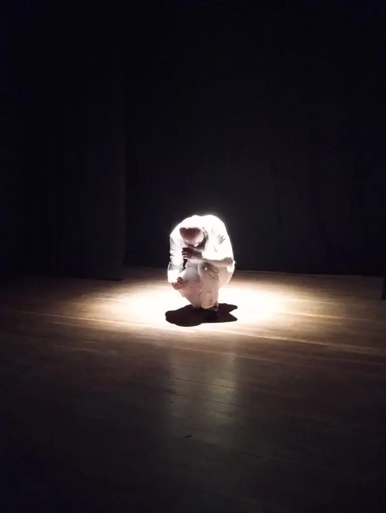 Sous un projecteur, une personne vêtue de blanc s'accroupit sur la scène, transmettant un sentiment d'introspection ou de réflexion dans un environnement sombre.