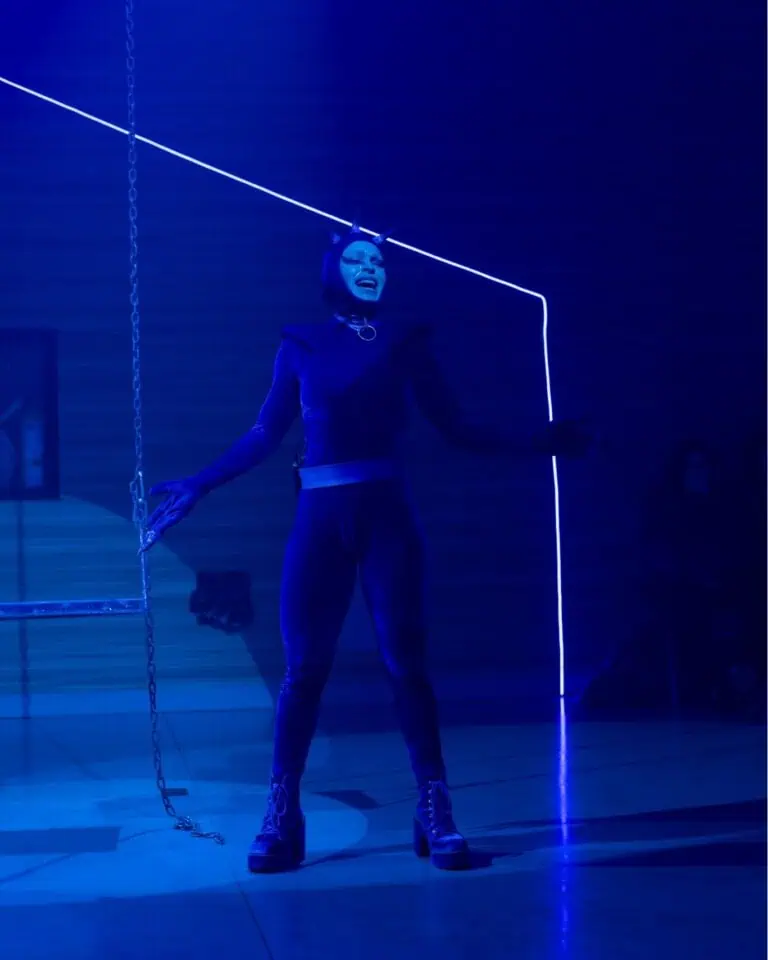 一位身着派对服装的表演者站在蓝色灯光下的舞台上，穿着奇幻的服装，佩戴着创意配饰，参加一场以艺术体验为重点的演出和娱乐活动。