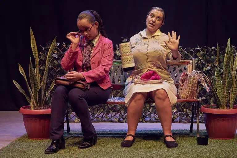 Personen in einem Theaterstück, eine davon mit überraschtem Gesichtsausdruck, sitzen auf einer Bank, umgeben von dekorativen Pflanzen, und spielen eine emotionale und komische Theaterszene.