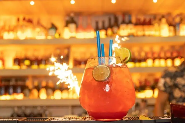 Colorful cocktail glass with lemons, several blue straws, and a light centerpiece, in a lively bar with bottle racks in the background.