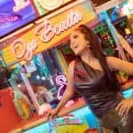 Oye Bonita, un stand de nourriture dans une foire avec un décor néon coloré, une femme souriante dans une robe en cuir et un pantalon métallique, une ambiance festive avec des lumières et des gens qui s'amusent.