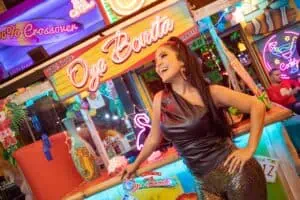 Oye Bonita, a food stand at a fair with colorful neon decor, a smiling woman in a leather dress and metallic pants, a festive atmosphere with lights and people enjoying themselves.
