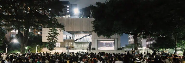 Une foule de personnes profitant d'un événement en plein air dans un cadre urbain la nuit, avec des arbres et des bâtiments modernes en arrière-plan.